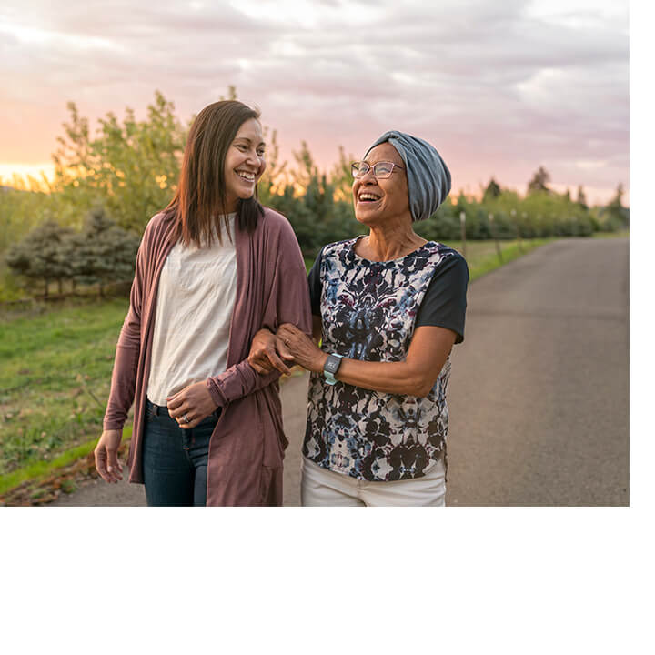 Two women, one a cancer survivor wearing a headscarf and her companion, walk outdoors at sunset, smiling and sharing a supportive moment