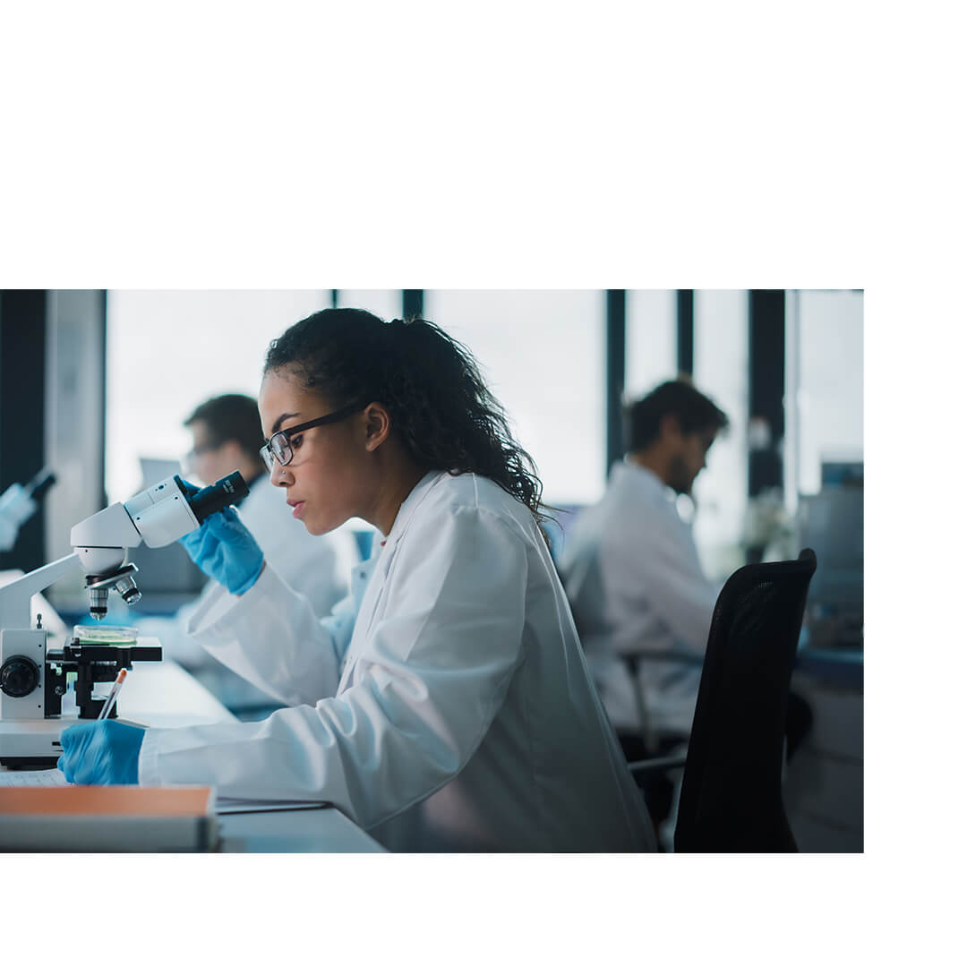 Female scientist wearing safety goggles and gloves operates a microscope in a cancer research lab