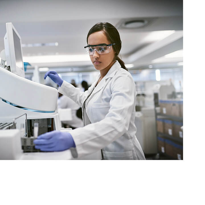 A female scientist in a lab coat and protective eyewear operates complex lab equipment in a bright research lab focused on cancer clinical trials