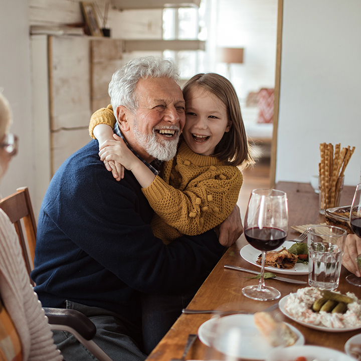 A young girl hugging her grandfather at the dinner table, both laughing and sharing a joyful moment