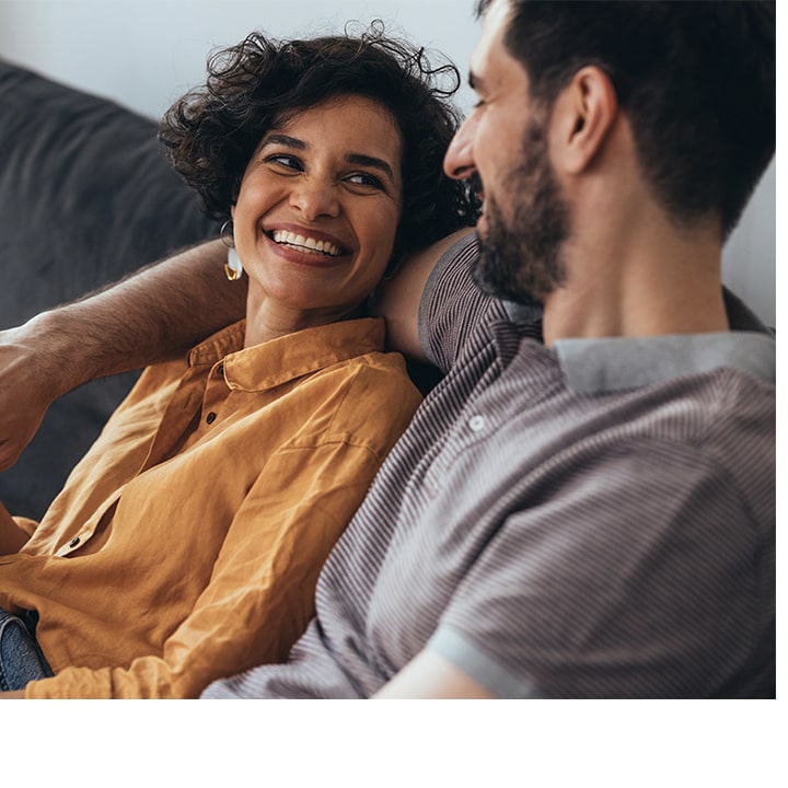 A smiling couple sits on a couch, discussing cancer risk prevention in a warm, sunny living-room setting