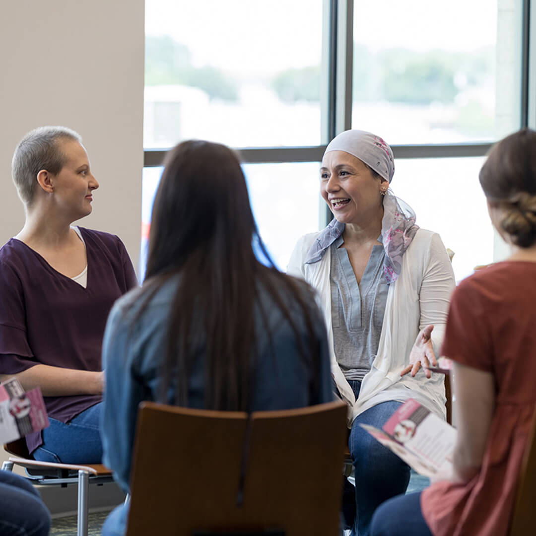 A group of adults sits in a cancer support group, in a well-lit room, looking comforted and engaged in conversation