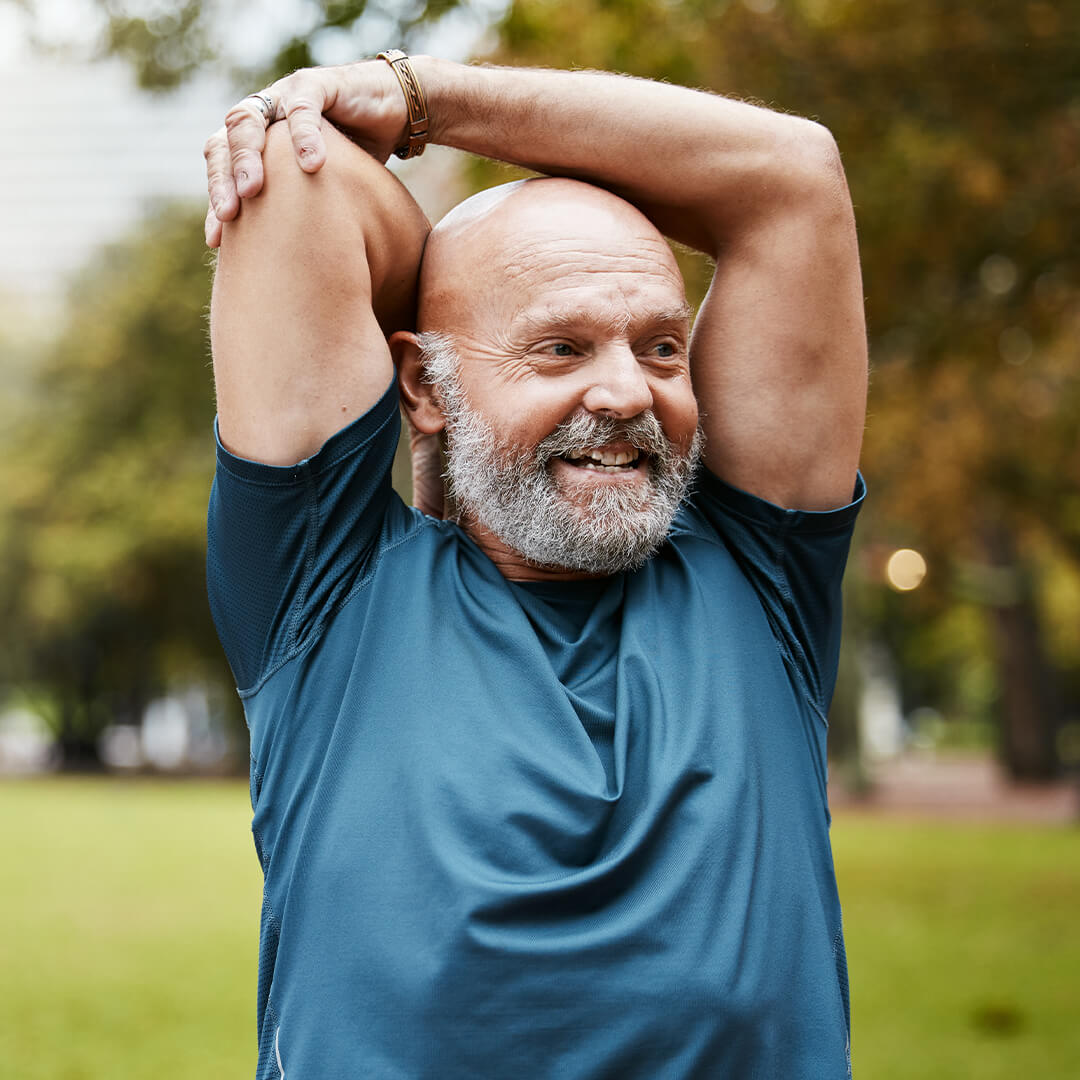 Bearded man in athletic wear stretches his arm above his head in an outdoor setting, taking part in a movement session for cancer survivors