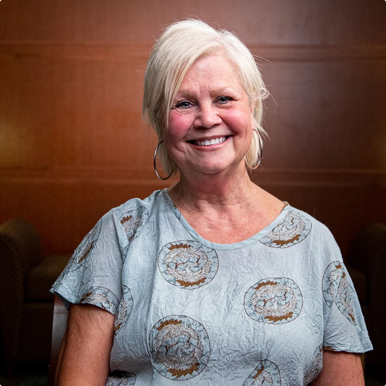Cindy Lesley, smiling in casual attire, sits against a neutral background, conveying warmth and hope as a cancer survivor