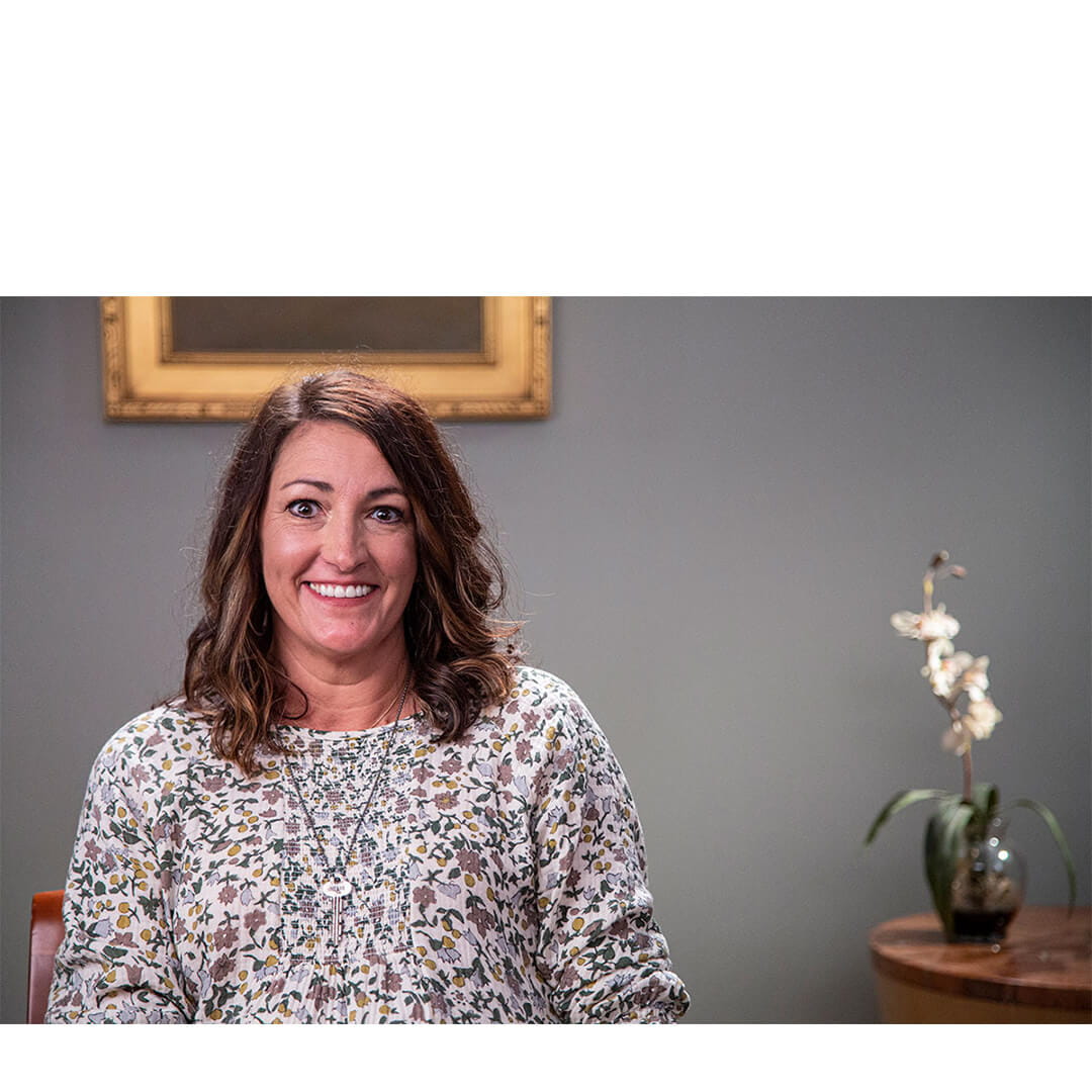 Ashleigh Parker, a smiling woman in casual attire, sits in a softly lit consultation room as she shares her cancer survivorship journey