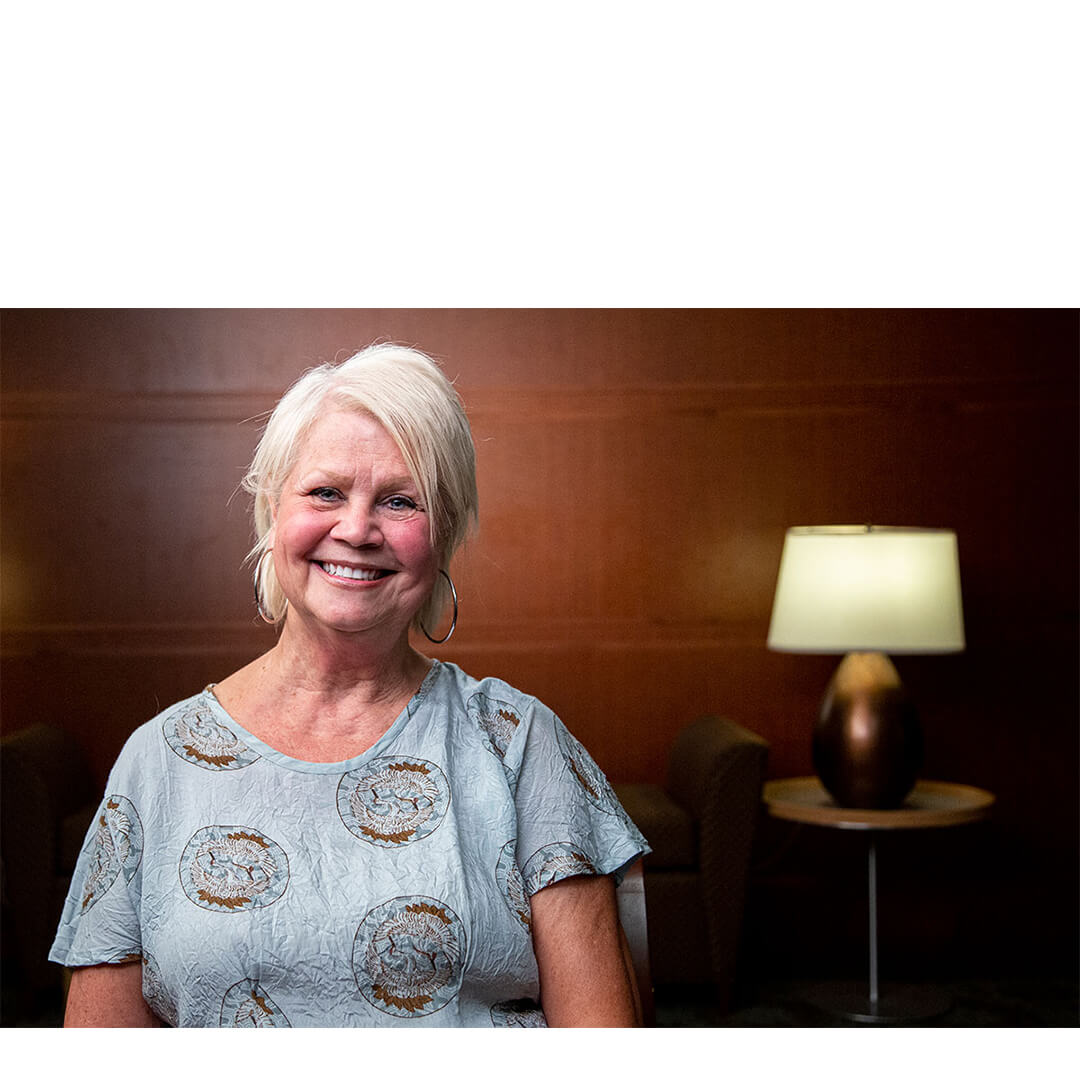 Cindy Lesley, smiling in casual attire, sits against a neutral background, conveying warmth and hope as a cancer survivor