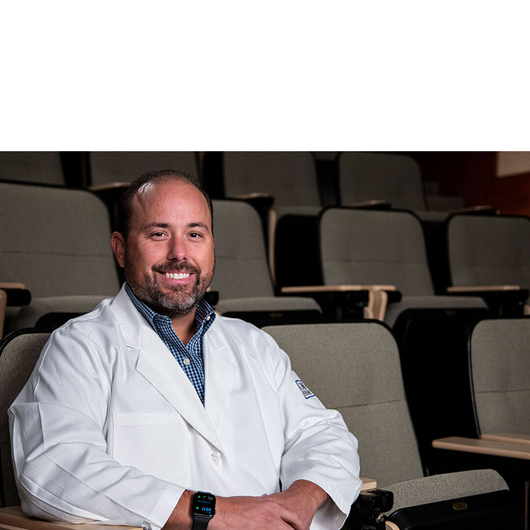 Dr. Drew Dill, a smiling hematology-oncology specialist in a white coat, sits confidently in an auditorium-style lecture hall