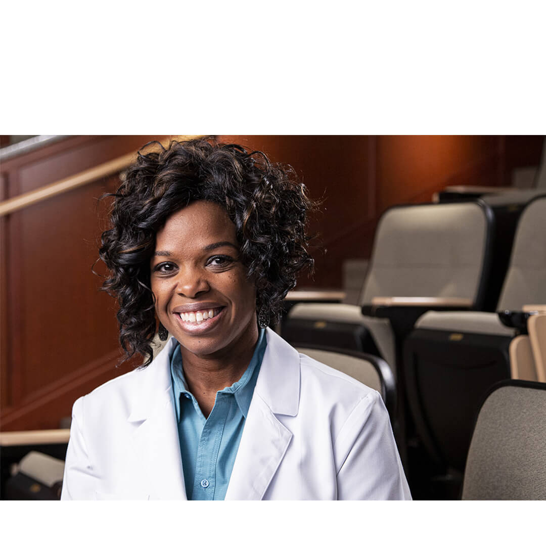 Clesheree Stepter, a smiling nurse practitioner in a white coat, sits in an auditorium-style clinic room, projecting confidence and compassion